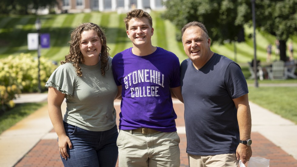 Two family members standing with their student in front of Donahue Hall.