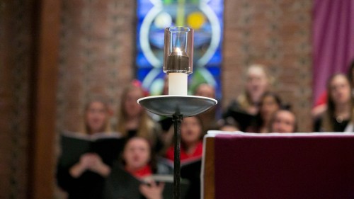 Candle in Chapel of Mary; Choir members are out of focus behind candle.