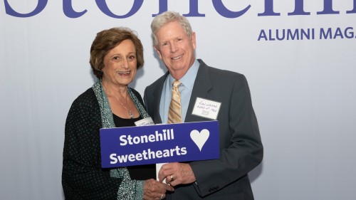 Couple in front of SAM masthead with sign that reads "Stonehill Sweethearts."
