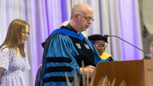 Fr. Adam Booth at podium in academic robes.