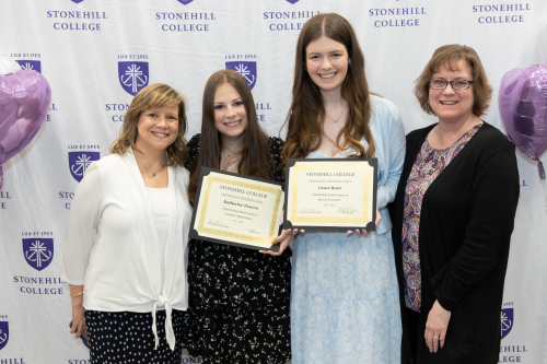 Four people are smiling and standing together, with two of them holding certificates from Stonehill College.