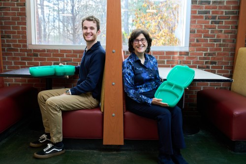 Caleb Hadley and Cheryl Schnitzer seated at booths in the Dining Commons, holding plastic green food containers