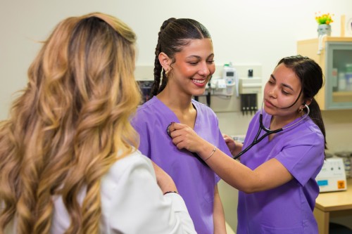 Two nursing students in purple scrubs, one listens to another's breathing using stethoscope.
