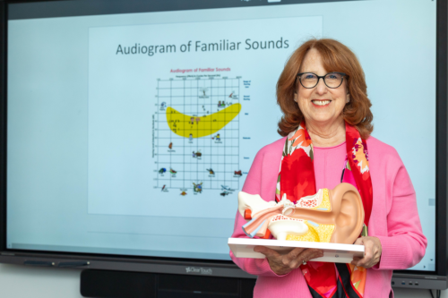 Robin Goldberg holding a model of the ear placed on table. Behind her is a white board with a diagram labeled "Audiogram of Familiar Sounds."