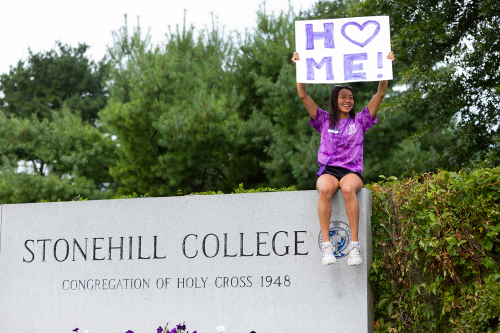 Peer mentor sitting on Stonehill sign, holding a sign that says "Home"