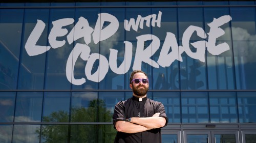 Fr. Tim Mouton in front of Sports Complex signage that says, "Lead with courage."