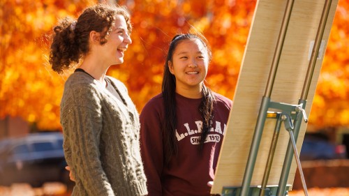Two students looking at board, fall leaves behind them.