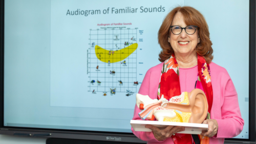 Robin Goldberg holding a model of the ear placed on table. Behind her is a white board with a diagram labeled "Audiogram of Familiar Sounds."