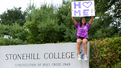 Peer mentor sitting on Stonehill sign, holding a sign that says "Home"